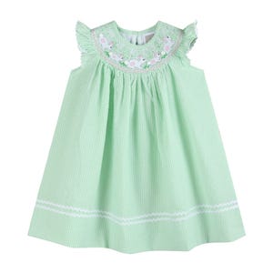May include: A light green seersucker dress with white stripes. The dress features a round neckline with embroidered bunnies and pink flowers. The sleeves have ruffled edges, and the hemline is trimmed with white scalloped detailing. A charming spring or Easter dress.