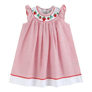 May include: A sleeveless dress featuring a red and white pinstriped seersucker fabric with a white yoke. The yoke is embroidered with red strawberries and green leaves. The dress has ruffled shoulders and a wide white hem with red trim.