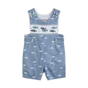May include: A light blue sleeveless romper featuring a white smocked chest panel with embroidered green and grey sharks. The garment is covered in a repeating pattern of small white shark outlines and has two decorative tabs with buttons at the waist.