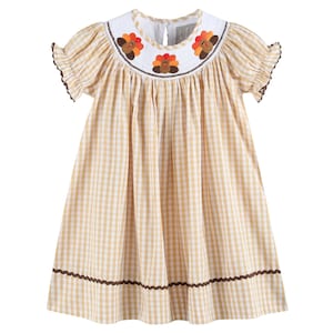 May include: A light yellow and white checkered dress with short sleeves and a white collar. The collar is embroidered with three turkey designs in brown, orange, and red. The dress has a scalloped brown trim at the sleeves and hem.