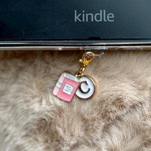 May include: A gold charm with a pink book and a white letter C attached to a black Kindle e-reader.