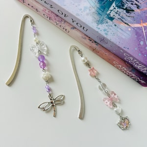 May include: Two silver bookmark with dangling charms. One bookmark has a dragonfly charm and the other has a butterfly charm. Both charms are silver with purple and pink accents. The bookmarks are on top of two books with the titles "Shadows of You" and "The Lost & Coveted".
