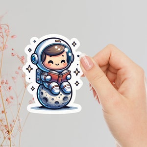 May include: A cartoon illustration of an astronaut wearing a white helmet and spacesuit sitting on the moon and reading a book. The astronaut has brown hair and is smiling. The moon is gray with craters. There are stars in the background.