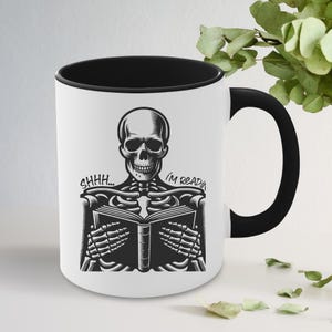 May include: White ceramic mug with a black interior and handle. The mug features a black and white illustration of a skeleton reading a book, with the text "SHHH... I'M READIN".