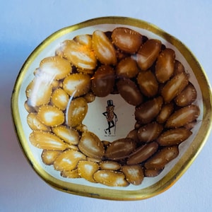 May include: A vintage metal dish with a gold rim and a white interior. The dish is filled with peanuts arranged in a circular pattern. The center features a cartoon image of Mr. Peanut. A classic collectible.