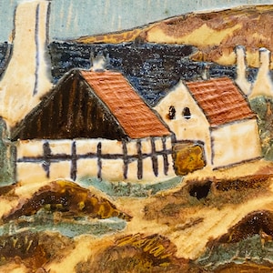 May include: A ceramic tile depicting a coastal village scene. The artwork features buildings with red tile roofs, set against a blue sky and sea. The houses have white walls and dark brown accents, with a textured, earthy landscape.