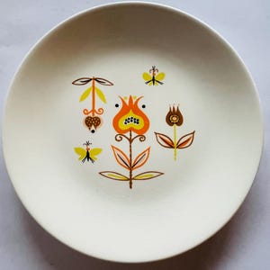 May include: A cream-colored ceramic plate with a floral design. The design features orange, yellow, and brown stylized flowers and butterflies. The plate has a slightly raised edge and a smooth surface.