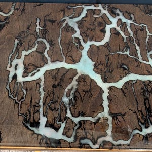 May include: A rectangular wooden table top with a unique design. The surface is a dark brown color with intricate white and black patterns resembling lightning or veins. The design is highlighted with a light blue-green resin.