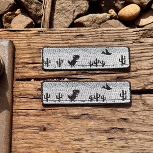 May include: Two rectangular patches with a grey background featuring a black dinosaur, birds, and cacti. The patches are on a wooden surface with rocks in the background.