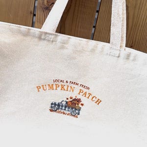 Peut inclure: Sac fourre-tout crème avec des anses blanches. Le sac présente un motif brodé d'un camion vintage transportant des citrouilles, avec les mots "Pumpkin Patch" et "Local & Farm Fresh" en orange.