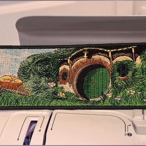 Könnte beinhalten: Gestickter Aufnäher mit einer detaillierten Darstellung eines Hobbit-Hauses mit grüner Tür, eingebettet in einen grasbewachsenen Hügel. Der Aufnäher hat einen schwarzen Rand und zeigt eine skurrile Landschaft mit lebendigen Farben.