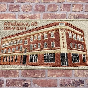 Peut inclure: Écusson brodé représentant un bâtiment en briques à plusieurs étages avec le texte "Athabasca, AB 1914-2024". Le bâtiment comporte de nombreuses fenêtres et une enseigne "Hotel". L'écusson est de forme rectangulaire.