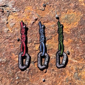 Könnte beinhalten: Drei gestickte Aufnäher von Kletterkarabinern mit farbigen Seilakzenten. Die Karabiner sind grau mit schwarzen Nähten, und die Seile sind rot, blau und grün. Die Aufnäher sind auf einer strukturierten, rostbraunen Oberfläche.