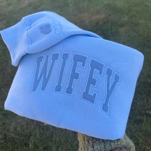 May include: A pale blue hoodie featuring the word "WIFEY" in raised lettering. The hood displays a small embroidered design and the text "EST.23". The sweatshirt is made of a soft material.