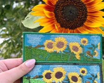 Pode incluir: Dois patches bordados com uma cena de campo de girassóis com girassóis amarelos, relva verde e céu azul. Os patches são retangulares com uma borda verde. Um grande girassol está no fundo.