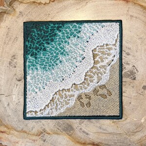 Peut inclure: Écusson carré brodé représentant une scène de plage. Le motif représente des vagues océaniques dans des tons de turquoise et de blanc, se jetant sur une plage de sable. Le patch a une bordure vert foncé et comprend des empreintes de pas.
