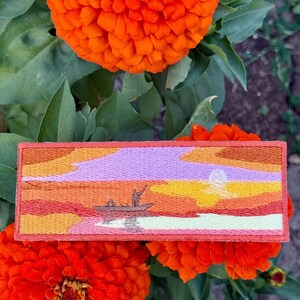 May include: Embroidered patch depicting a sunset scene with a boat and figures, in shades of orange, yellow, and purple. The rectangular patch has a red border, and is set against a backdrop of orange flowers and green foliage.