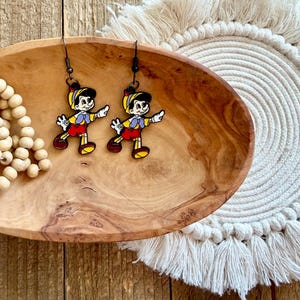 May include: A pair of dangle earrings featuring a cartoon character in yellow, red, and blue. The earrings are displayed on a wooden oval dish, next to a strand of wooden beads and a white macrame coaster. The background is a wooden surface.