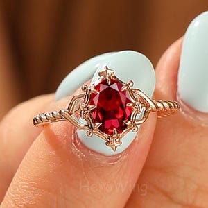 May include: A rose gold ring featuring a large, oval-cut, deep red gemstone. The gemstone is set in a decorative filigree setting with a twisted band. The ring is held up by a hand with light blue nail polish.