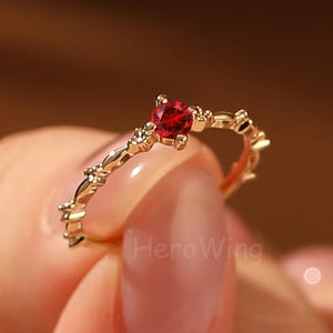 May include: A gold-colored ring with a heart-shaped, deep red gemstone at the center. The band features delicate, decorative accents. The ring is held up by a person's fingers.