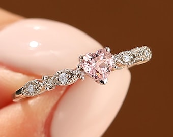 May include: A delicate silver ring featuring a heart-shaped pink gemstone as the centerpiece. The band is adorned with small, sparkling accent stones. The ring is held up against a neutral background, showcasing its elegant design.