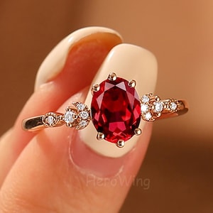 May include: A gold ring with a large, oval-cut, deep red gemstone as the centerpiece. The ring is decorated with small, clear accent stones on either side of the main stone. The ring is held up by a person's hand.