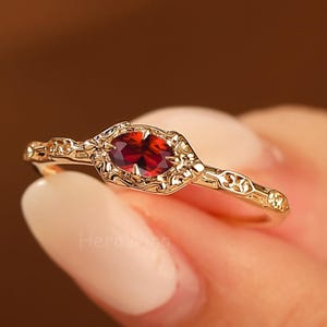 Könnte beinhalten: Ein goldfarbener Ring mit einem ovalen, tiefroten Edelstein. Der Ring hat ein verziertes Filigranmuster um den Edelstein und entlang des Rings. Der Ring wird vor einem unscharfen Hintergrund gehalten.