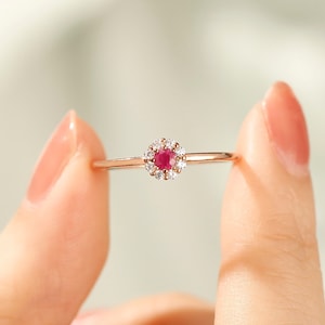 K&ouml;nnte beinhalten: Ein Ring aus Ros&eacute;gold mit einem roten Rubin-Edelstein, der von einem Heiligenschein aus kleineren wei&szlig;en Edelsteinen umgeben ist.