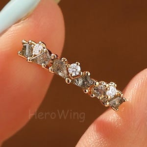 May include: A gold ring with a series of small, irregularly shaped gray and clear stones. The ring is held between two fingers with light blue nail polish. The brand name "HeroWing" is visible on the ring.