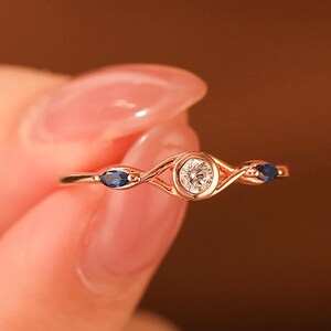 May include: A delicate gold ring featuring a central, round, clear gemstone, flanked by two oval, dark blue gemstones. The ring has a slender band with a decorative, openwork design. The ring is held up by a person's fingers.