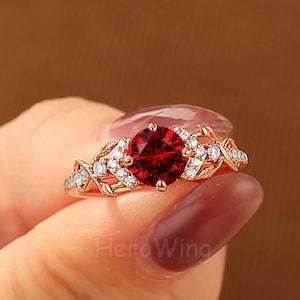 May include: A rose gold ring featuring a round, deep red center stone flanked by small, clear accent stones. The band is crafted with an intricate, openwork leaf and vine design that extends along the sides of the central setting.