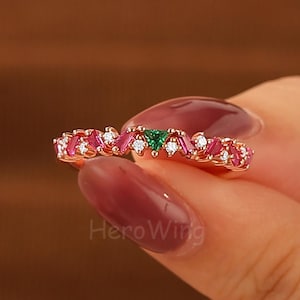 May include: A rose gold ring featuring alternating pink and white gemstones, with a central green gemstone. The ring is held up by a person's hand. The brand name "Hero Wing" is visible on the hand.