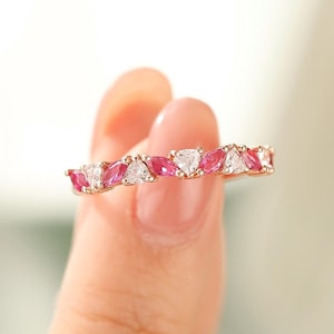 May include: A gold-toned ring featuring alternating pink and white gemstones. The gemstones are cut into a unique shape, creating a sparkling and elegant design. The ring is held up by a person's fingers.