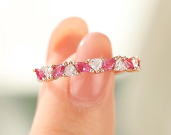 May include: A gold-toned ring featuring alternating pink and white gemstones. The gemstones are cut into a unique shape, creating a sparkling and elegant design. The ring is held up by a person's fingers.
