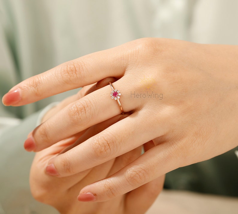 K&ouml;nnte beinhalten: Ein Ring aus Ros&eacute;gold mit einem roten Edelstein in der Mitte, umgeben von kleineren klaren Edelsteinen. Der Ring befindet sich an einem Finger.