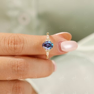May include: A gold ring featuring an oval-cut, color-changing gemstone with a deep blue and purple hue. The ring is accented with small, clear gemstones. The ring is held by a person.