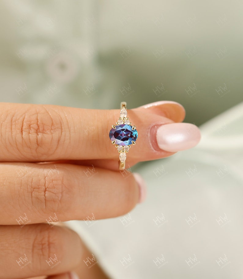 May include: A gold ring featuring an oval-cut, color-changing gemstone with a deep blue and purple hue. The ring is accented with small, clear gemstones. The ring is held by a person.