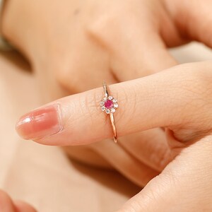 K&ouml;nnte beinhalten: Ein zarter Goldring mit einem kleinen roten Edelstein, der von einem Heiligenschein aus kleineren wei&szlig;en Edelsteinen umgeben ist.