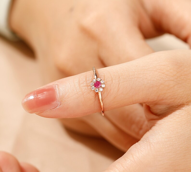 K&ouml;nnte beinhalten: Ein zarter Goldring mit einem kleinen roten Edelstein, der von einem Heiligenschein aus kleineren wei&szlig;en Edelsteinen umgeben ist.