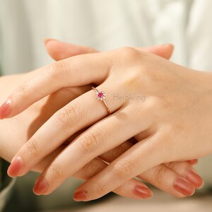 K&ouml;nnte beinhalten: Ein Ring aus Ros&eacute;gold mit einem kleinen roten Edelstein, der von kleineren wei&szlig;en Edelsteinen umgeben ist. Der Ring befindet sich an einem Finger.