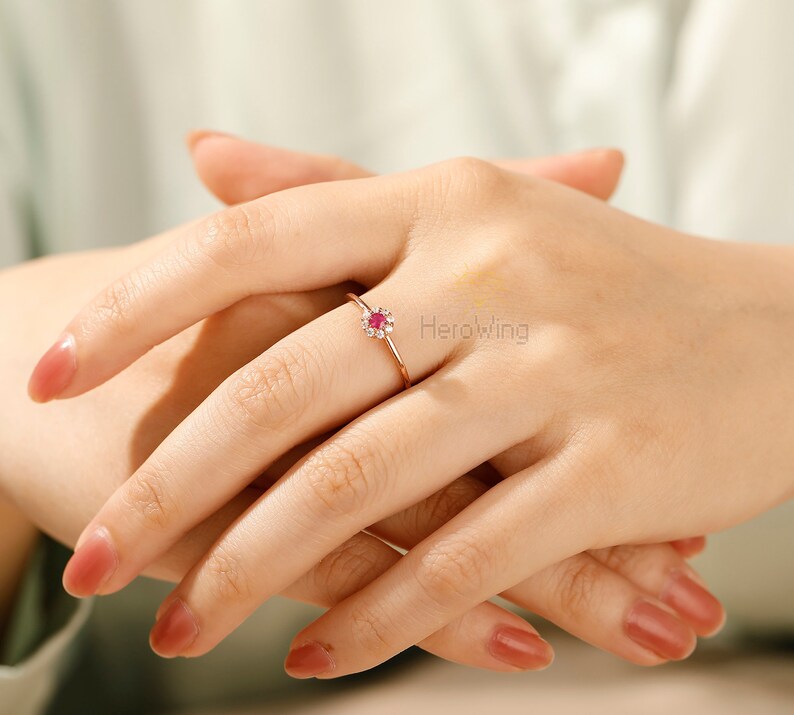 K&ouml;nnte beinhalten: Ein Ring aus Ros&eacute;gold mit einem kleinen roten Edelstein, der von kleineren wei&szlig;en Edelsteinen umgeben ist. Der Ring befindet sich an einem Finger.