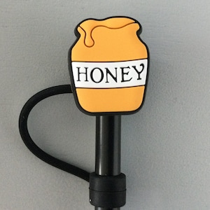 May include: A black and yellow honey pot shaped bottle stopper with the word "Honey" printed on the front. The bottle stopper is attached to a black cord.