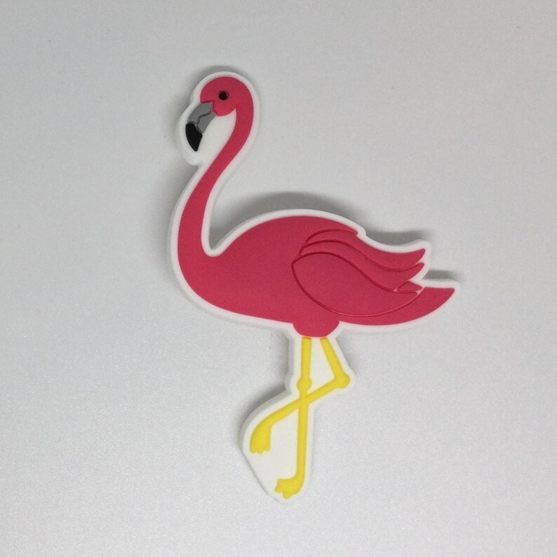 May include: A pink flamingo-shaped decorative item with a white outline. The flamingo has a black beak, a red eye, and yellow legs. The item is likely made of a rubbery material.