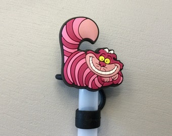 Op de afbeelding: Een roze en wit Cheshire Cat-personage uit Alice in Wonderland. De kat glimlacht met wijd open ogen. Het personage is gemaakt van rubber en is bevestigd aan een witte stok.