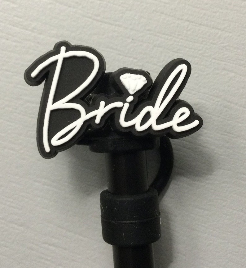 以下が含まれることがあります： ダイヤモンドアクセントが付いた、黒と白の「Bride」チャーム。結婚式をテーマにしたアクセサリー用の黒い茎に取り付けられています。「Bride」の文字は筆記体です。