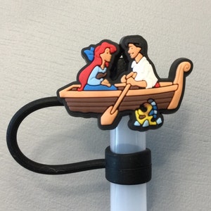 May include: A black plastic straw holder with a cartoon image of Ariel and Prince Eric from The Little Mermaid in a boat. The boat is brown with a red oar and a yellow fish.