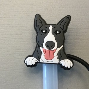 May include: A black and white bull terrier dog head peeking out from behind a white plastic pen holder. The dog has a pink tongue sticking out and a friendly expression.