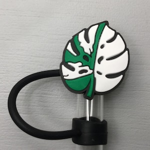May include: A black silicone straw holder with a green and white monstera leaf design. The holder has a loop for attaching to a cup or bottle.