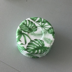 May include: A white round lid with a green tropical leaf pattern. The lid has a glossy finish.