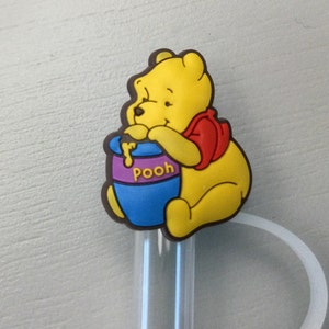 May include: Yellow Winnie the Pooh cartoon character holding a blue honey pot with the word "Pooh" on it. The character is a straw topper.